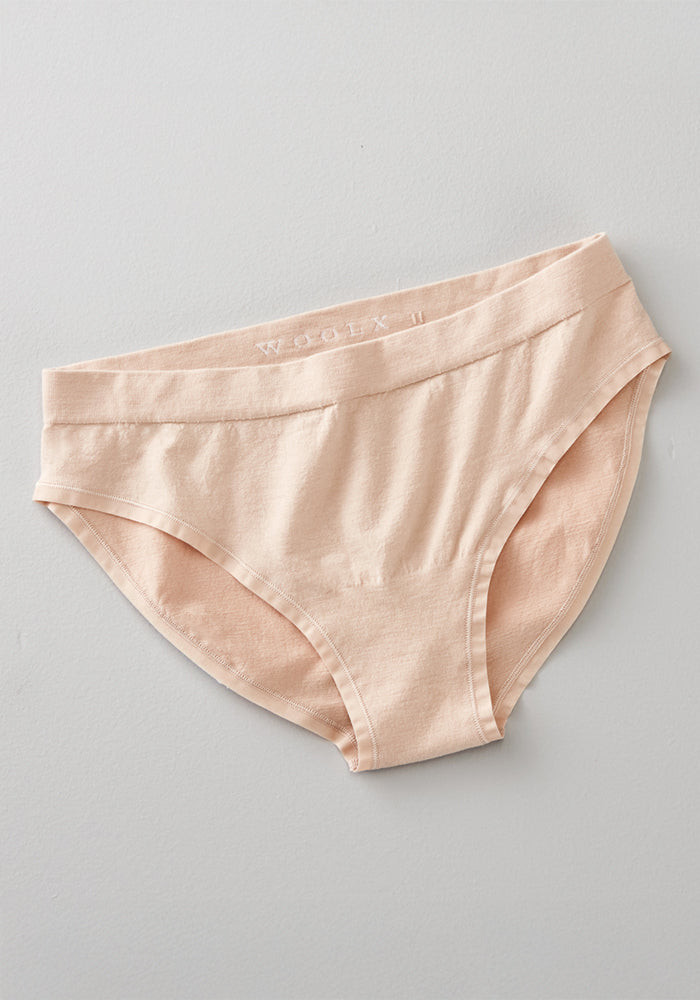 The Woolx Maeve Bikini in Sand is a seamless, light beige women’s underwear with a mid-rise waistband and moisture-wicking fabric, shown laid flat on a light grey surface.