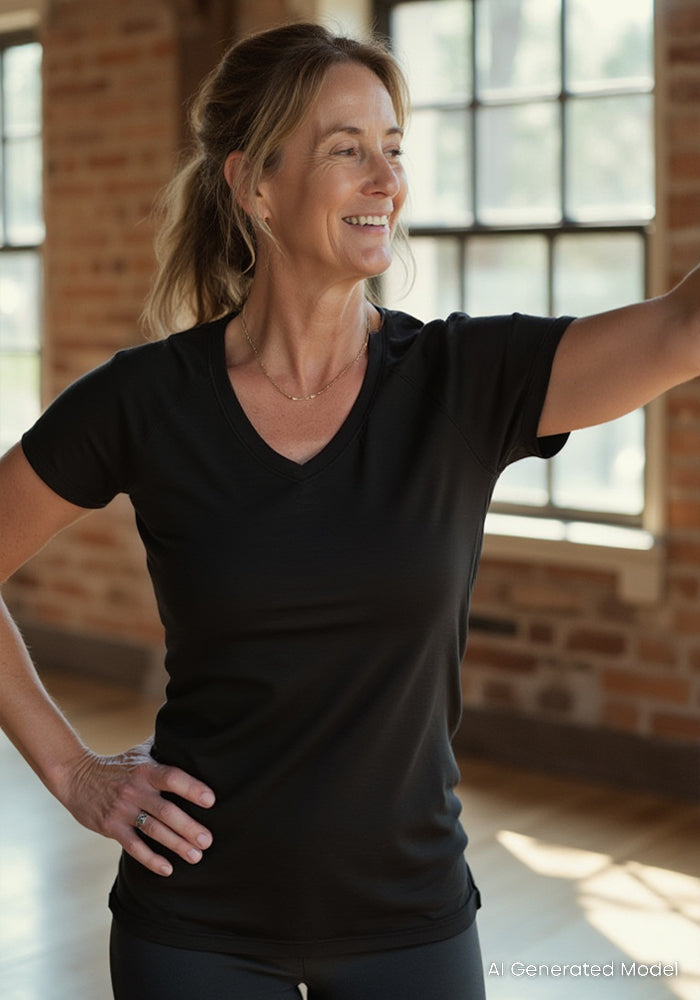 A woman smiles indoors with her hand on her hip, wearing the Woolx Mia V Neck in black. Sunlight streams through large windows onto the wooden floor and brick walls as she appears to wave or reach out.