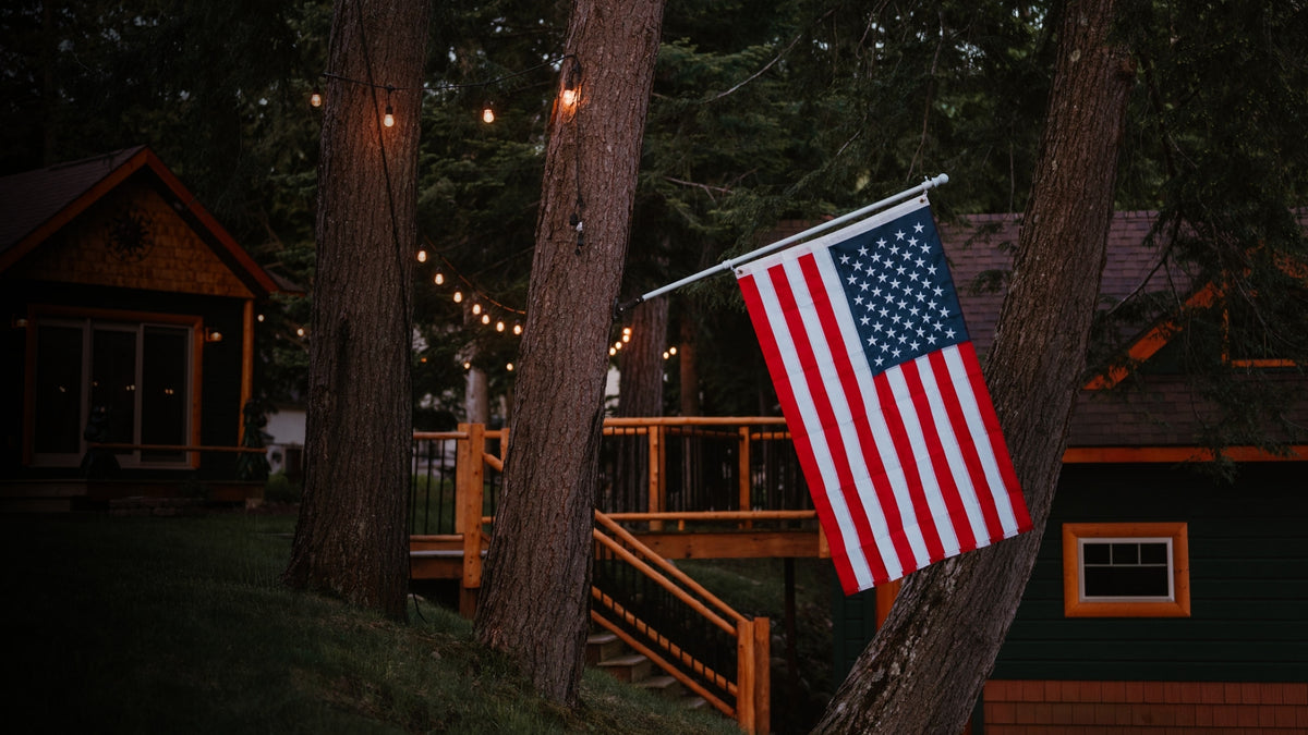 An American flag hanging from a metal pole mounted on a tree outside a cabin, with wooden railings and dark trees in the background.