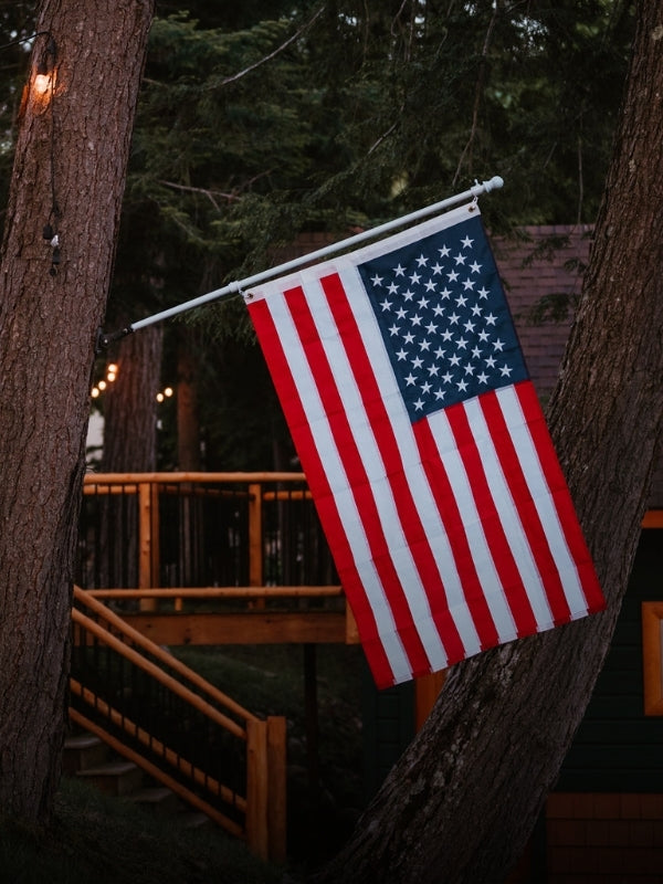 An American flag hanging from a metal pole mounted on a tree outside a cabin, with wooden railings and dark trees in the background.