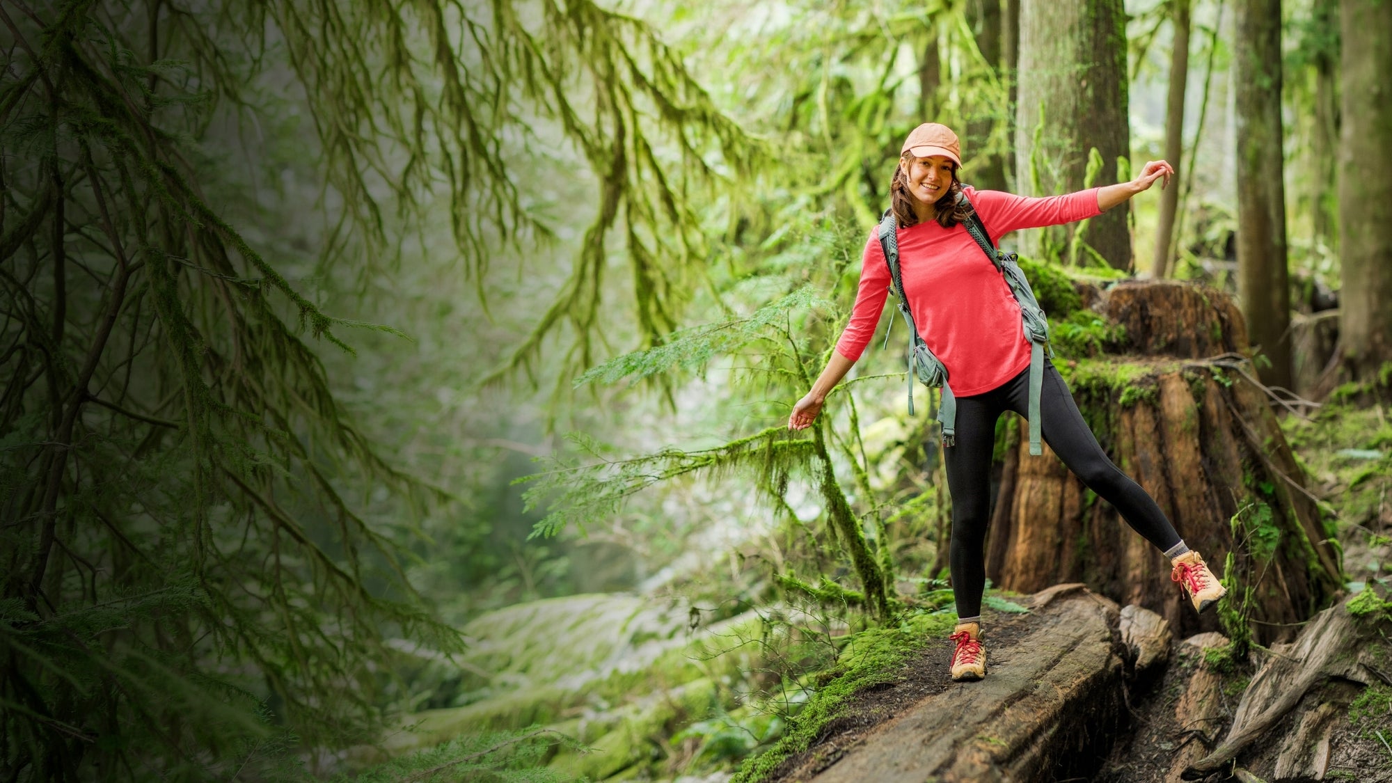 A woman hiking through a lush green forest, wearing a bright pink long-sleeve top, black leggings, and a pink cap, carrying a blue backpack.