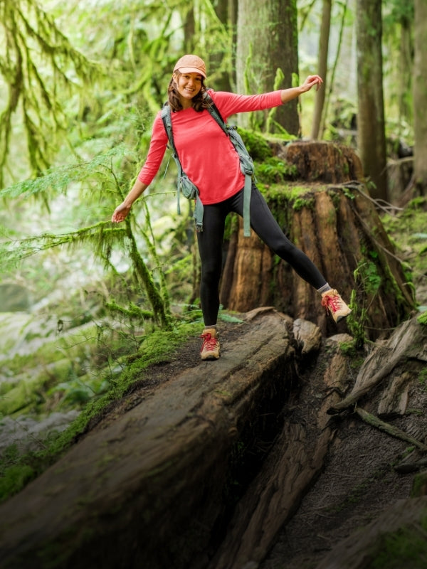 A woman hiking through a lush green forest, balancing on a large fallen log. She is wearing a coral pink long-sleeve top, black leggings, pink hiking shoes, and a gray backpack.