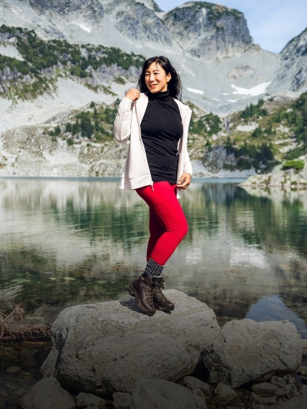A woman standing on the shore of a mountain side lake wearing bright red leggings and a zip up hoodie.