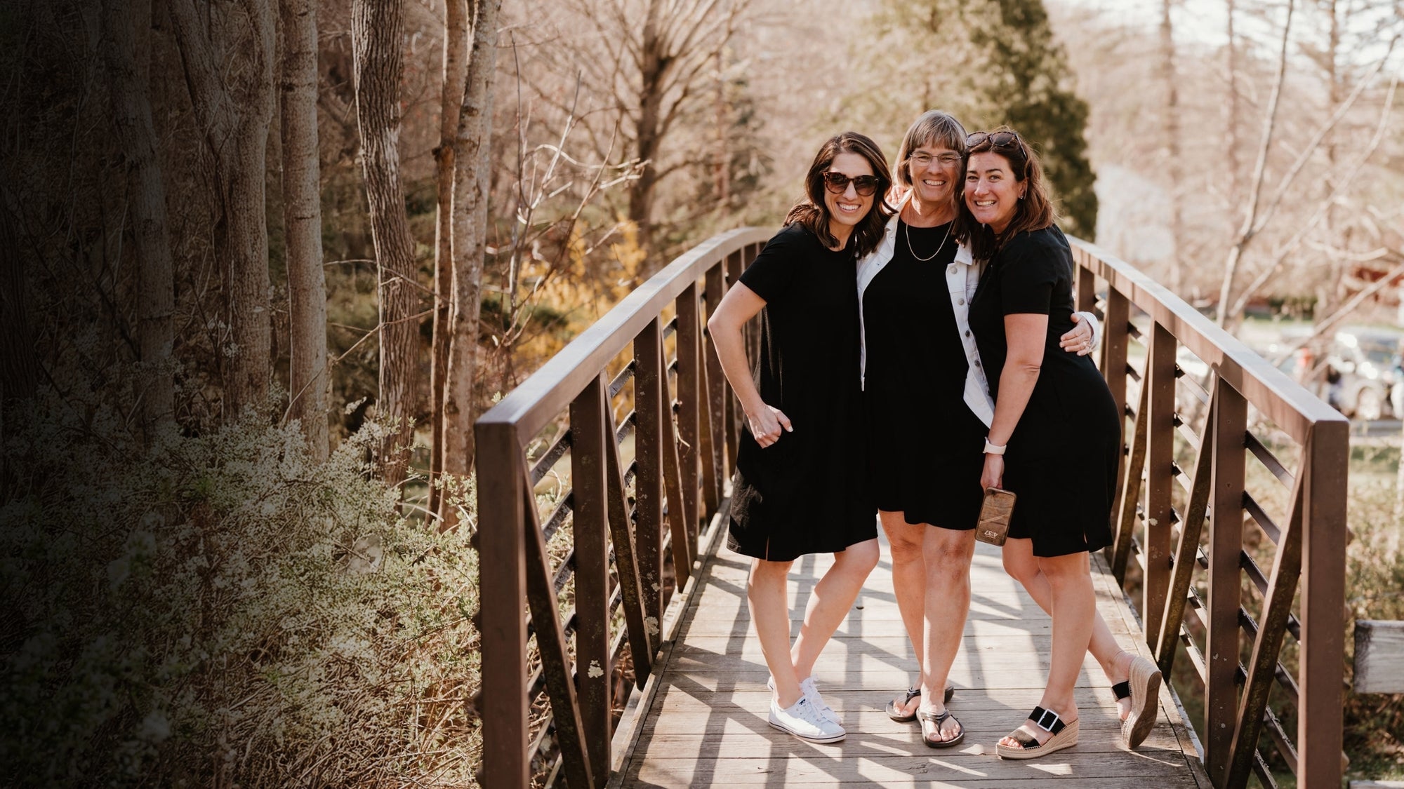 Three women wearing black standing on a wooden bridge in a wooded area posing for the camera. 