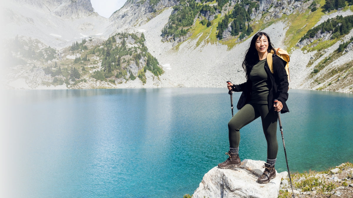 A woman hiking with trekking poles stands on a rock overlooking a bright blue alpine lake, with rocky mountains and green vegetation in the background.