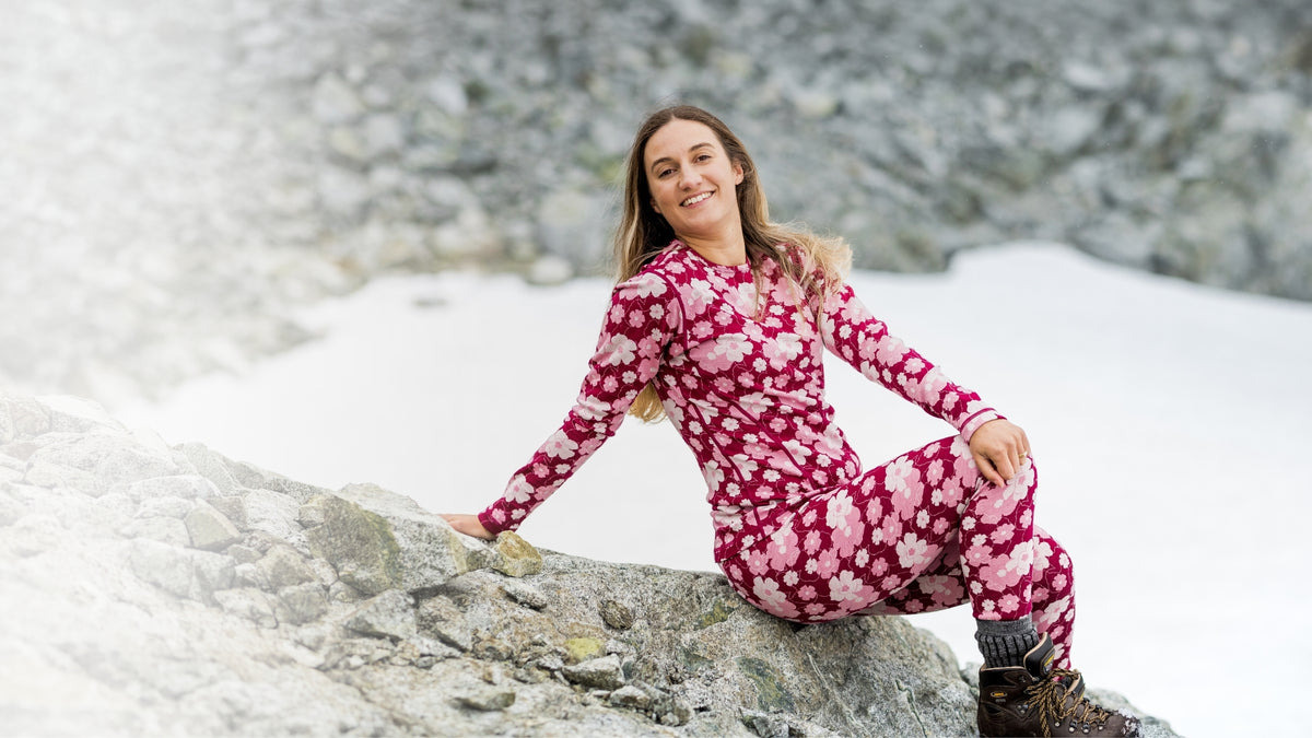 Woman sitting on a rock in a snowy landscape, wearing a burgundy merino base layer top and leggings with a pink floral print.