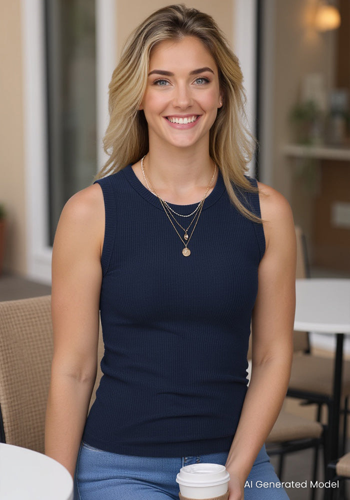 A smiling woman with wavy blonde hair wears the Woolx Raegan Ribbed Tank Top in Deep Navy and layered necklaces, sitting outdoors with a coffee cup. In the background are a beige building and blurred chairs.