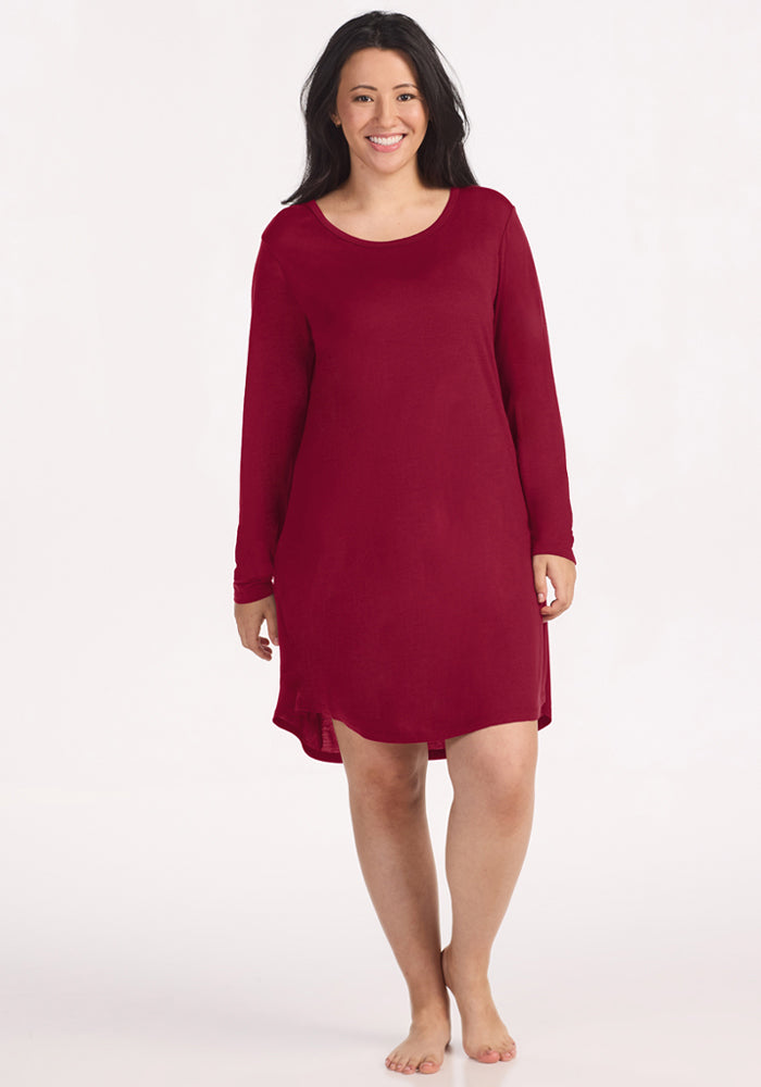 A smiling woman wears a beet red, long-sleeve Woolx Lucille Long Sleeve Nightgown with a rounded hem.
