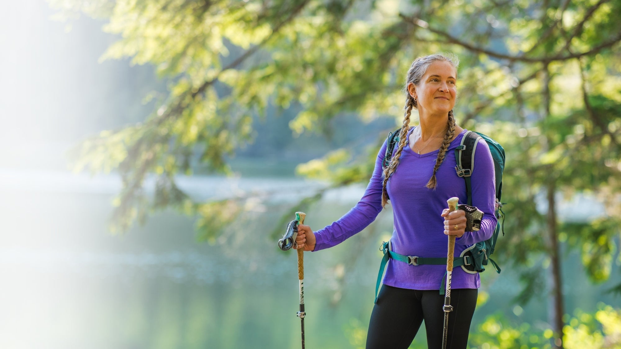 A woman hiking outdoors near a lake, wearing a purple long-sleeve top, black leggings, a green backpack, and holding hiking poles.
