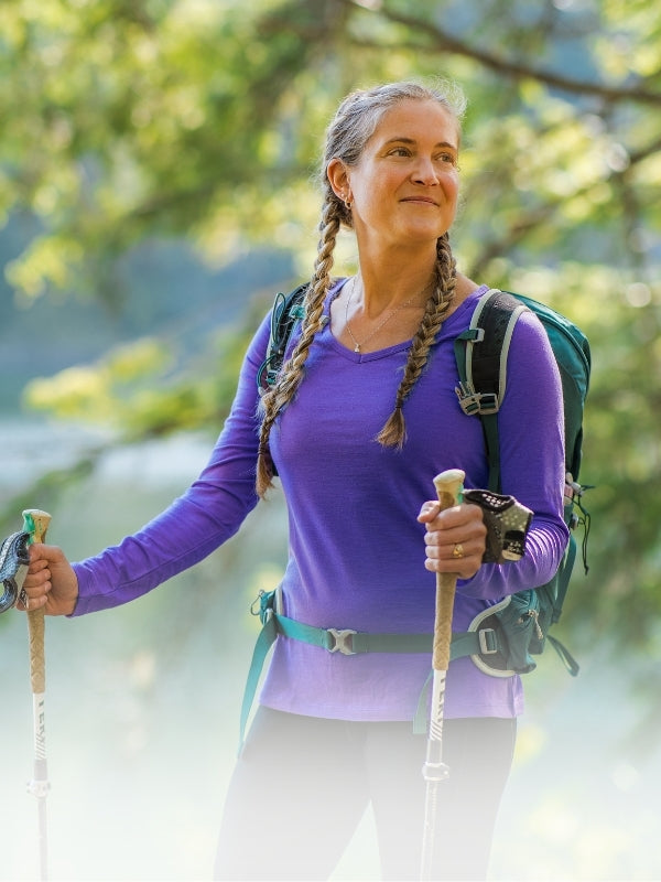 A woman hiking outdoors near a lake, wearing a purple long-sleeve top, black leggings, a green backpack, and holding hiking poles.