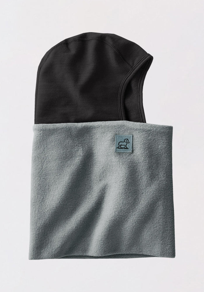 The Woolx Shellaclava - Sedona Sage is a two-tone, helmet-compatible hooded neck warmer featuring a black hood and light gray fleece for top cold-weather protection. A small blue patch with an embroidered logo accents the gray section in front.
