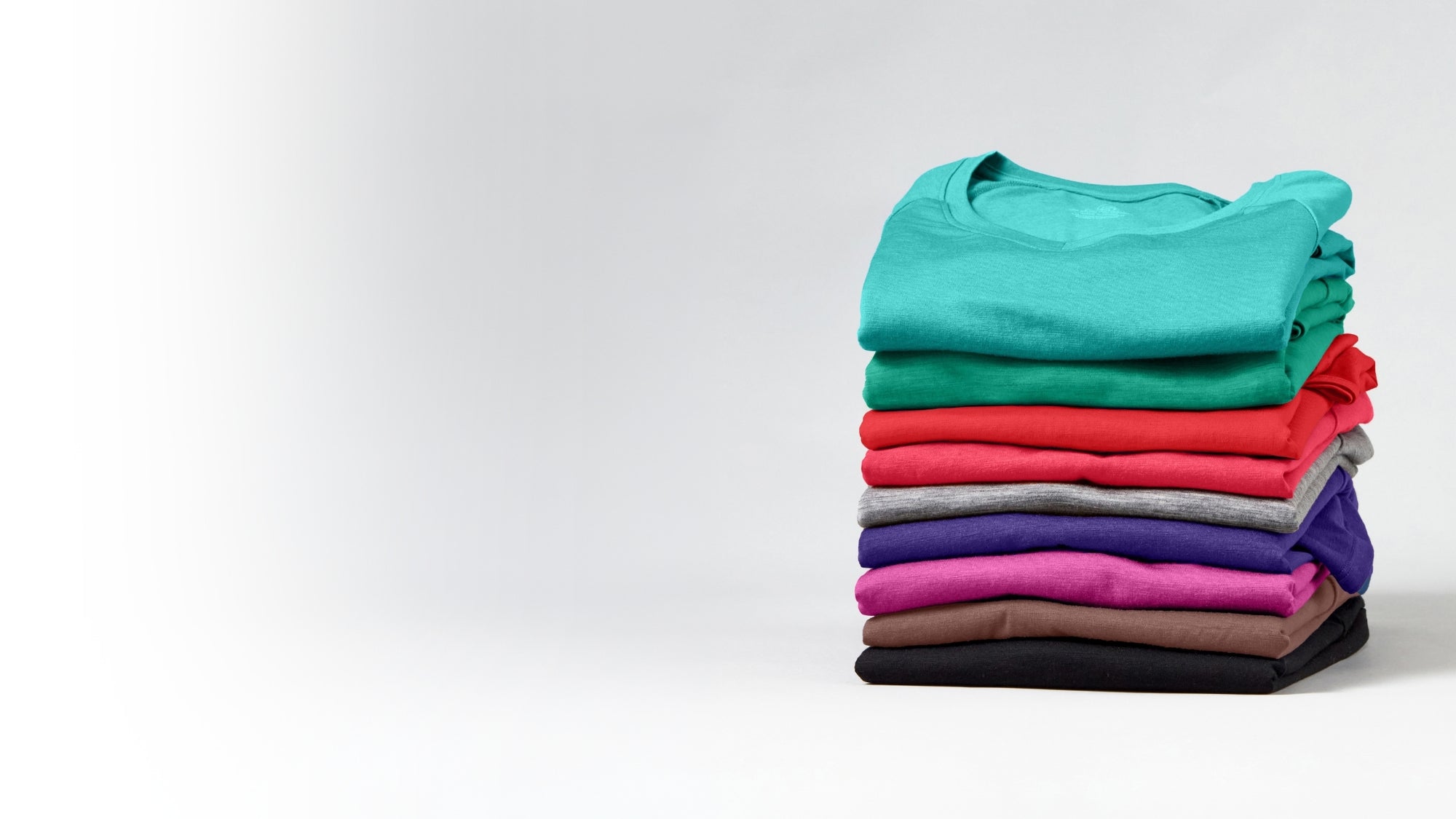 A stack of neatly folded crewneck t-shirts in a variety of colors, including teal, green, red, orange, gray, purple, pink, brown, and black, set against a clean white studio background.