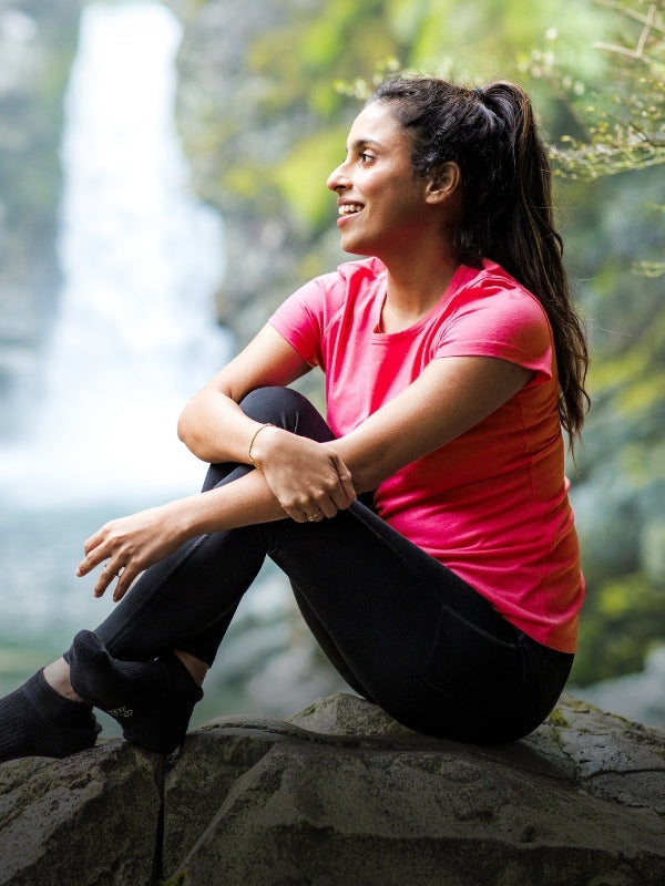 Woman in pink shirt and black leggings sitting on a rock with a blurred natural background