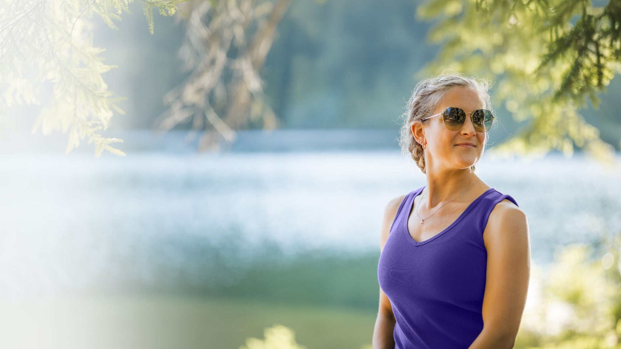 A woman wearing a purple sleeveless top and sunglasses stands outdoors near a blurred lake or river with trees in the background.