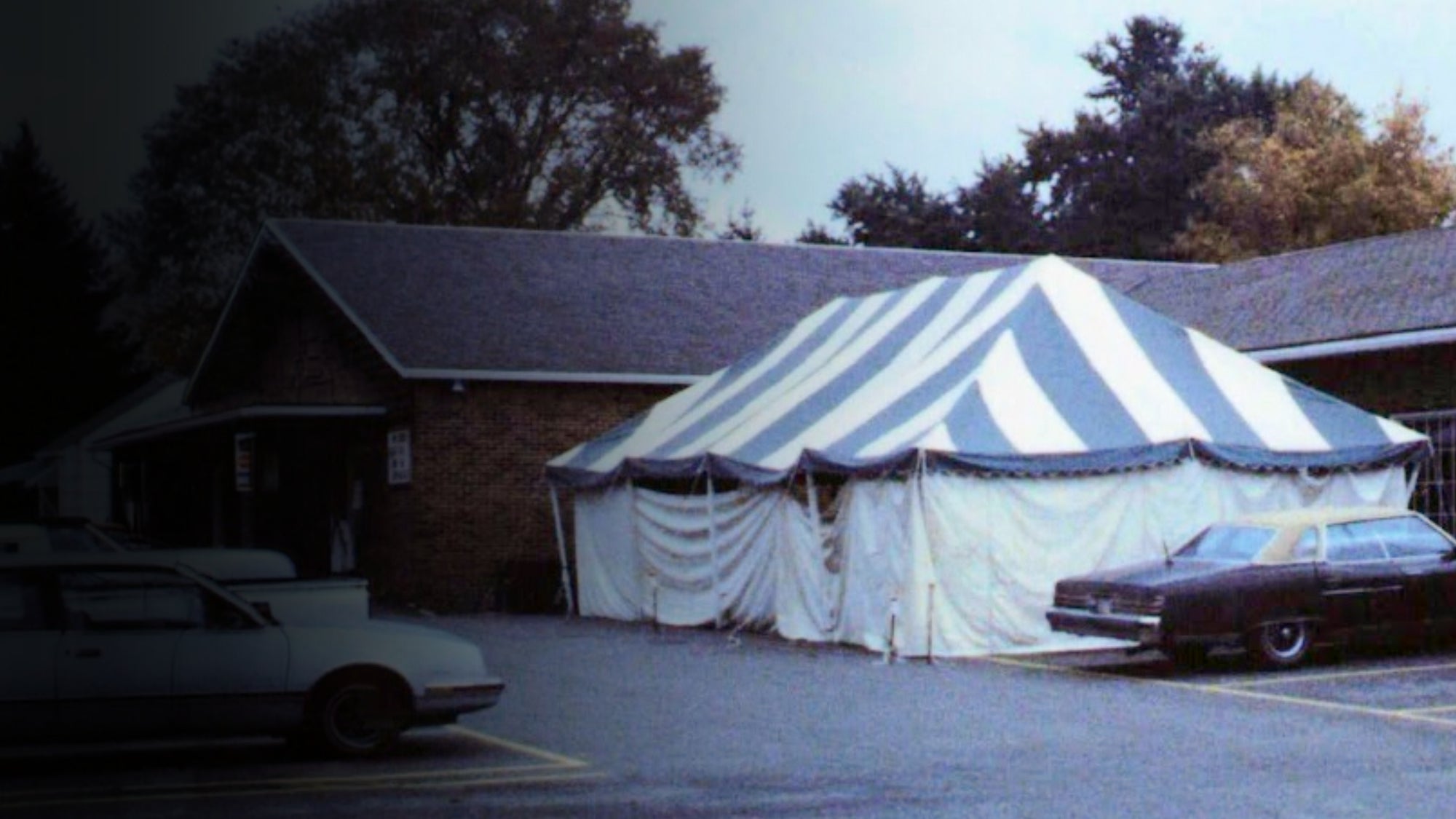 A large striped event tent with blue and white vertical stripes is pitched in a parking lot in front of a brick building, with two cars parked nearby.