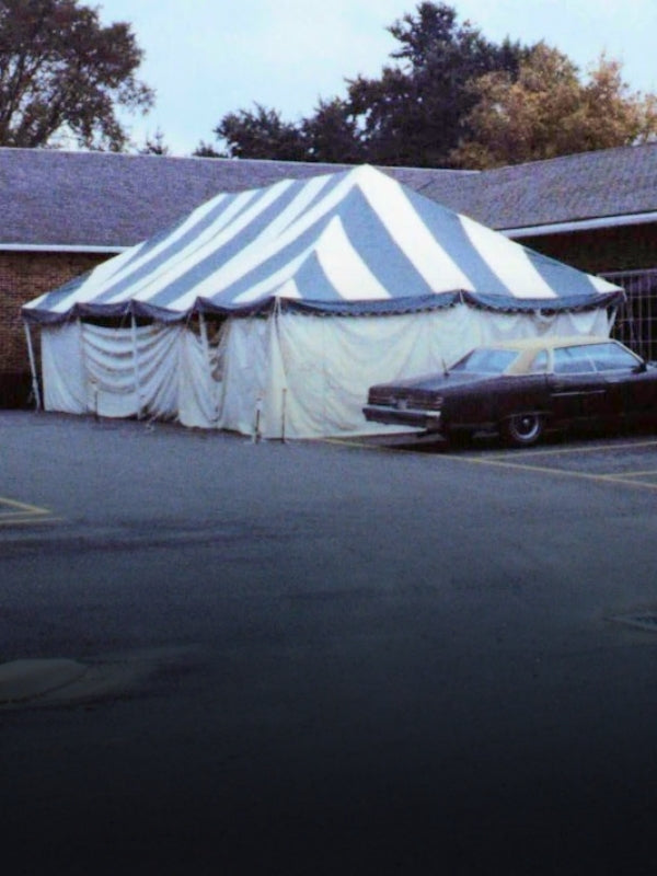 A large striped event tent with blue and white vertical stripes and white side curtains, pitched next to a dark car in an outdoor parking lot.