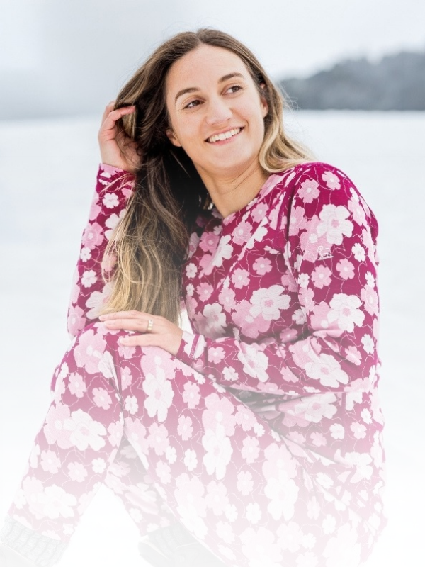 
Smiling woman seated in the snow, wearing a burgundy merino base layer top and leggings with a pink floral print.