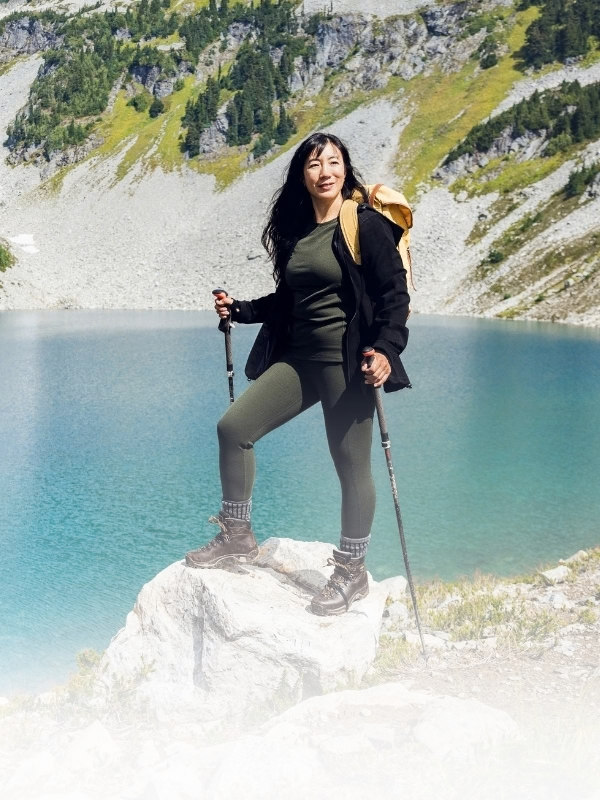A woman hiking with trekking poles stands on a rock overlooking a bright blue alpine lake, with rocky mountains and green vegetation in the background.