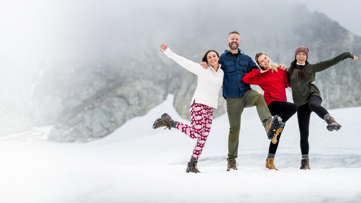 Four adults in colorful cold-weather outfits pose playfully in the snow against a mountain backdrop.