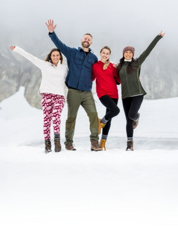 Four adults in colorful cold-weather outfits pose playfully in the snow against a mountain backdrop.