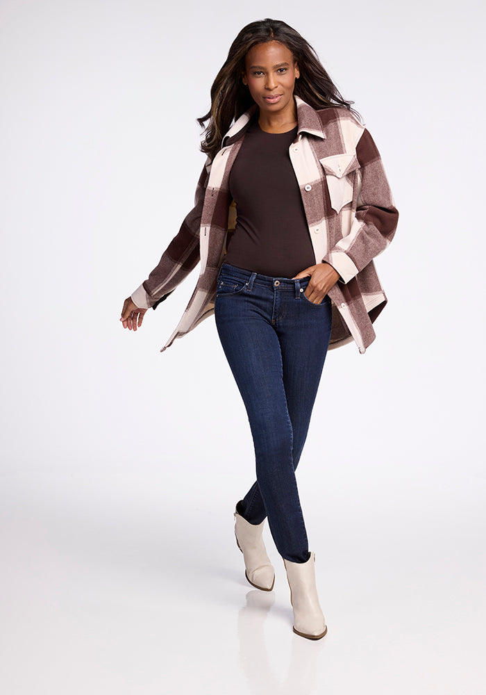 A woman with long dark hair walks confidently against a white background, wearing the Woolx Sawyer Shacket in Chai Latte Plaid over a dark top, blue jeans, and white ankle boots.