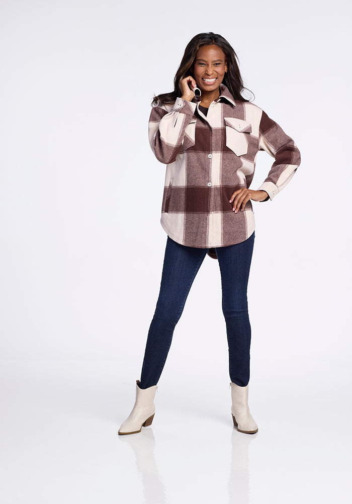 A woman smiles while talking on the phone, wearing the Woolx Sawyer Shacket in Chai Latte Plaid, paired with dark blue jeans and cream ankle boots. She stands confidently against a white background.