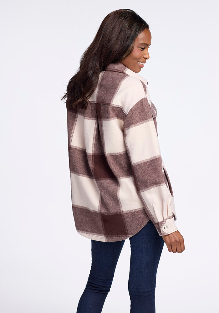 A woman with long dark hair wears the Woolx Sawyer Shacket in Chai Latte Plaid and dark blue jeans, standing sideways and smiling against a plain light background.