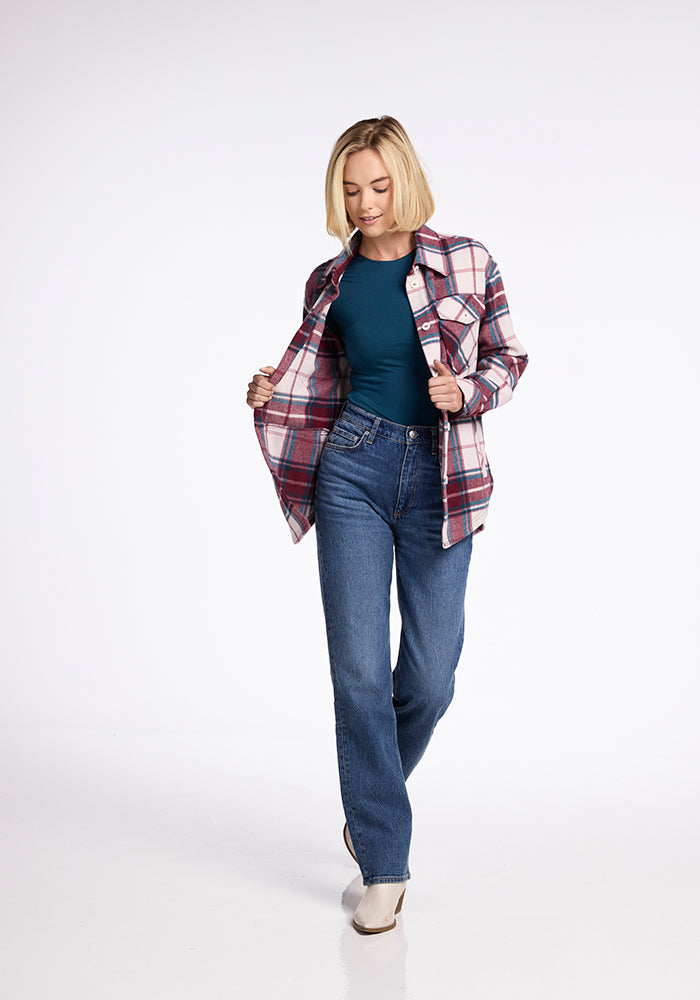A person with short blonde hair is wearing a Woolx Sawyer Shacket over a teal top and jeans. They are looking down and holding the plaid shacket open with both hands. The background is plain white.