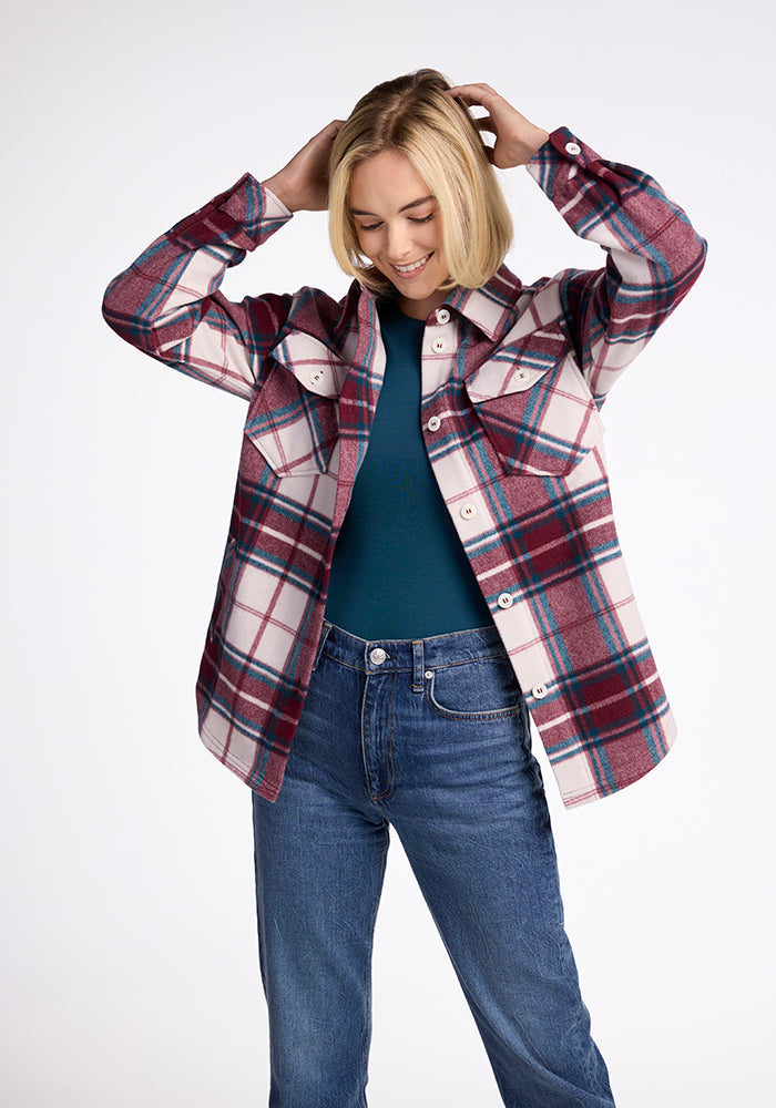 Someone wearing a Woolx Sawyer Shacket in Frozen Cranberry Plaid over a teal top and blue jeans stands against a plain background, looking down and smiling while adjusting their hair.