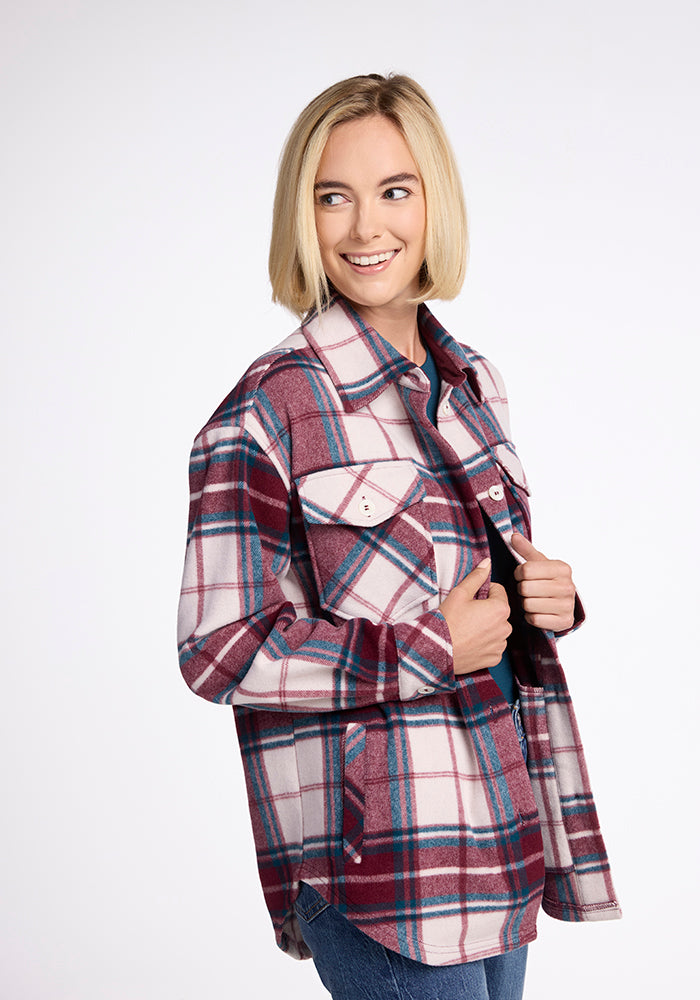 A person with short blonde hair smiles, wearing the Woolx Sawyer Shacket in Frozen Cranberry Plaid over a dark top, looking slightly to the side against a plain white background.