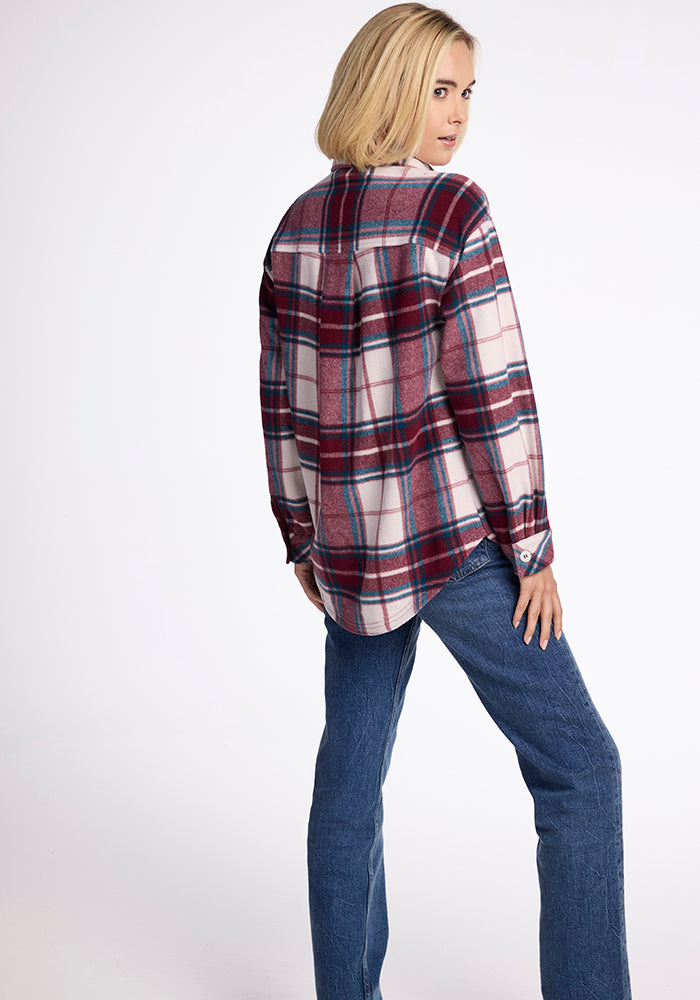 A person with shoulder-length blonde hair stands with their back to the camera, wearing Woolx's Sawyer Shacket in Frozen Cranberry Plaid and blue jeans, glancing slightly over their shoulder.