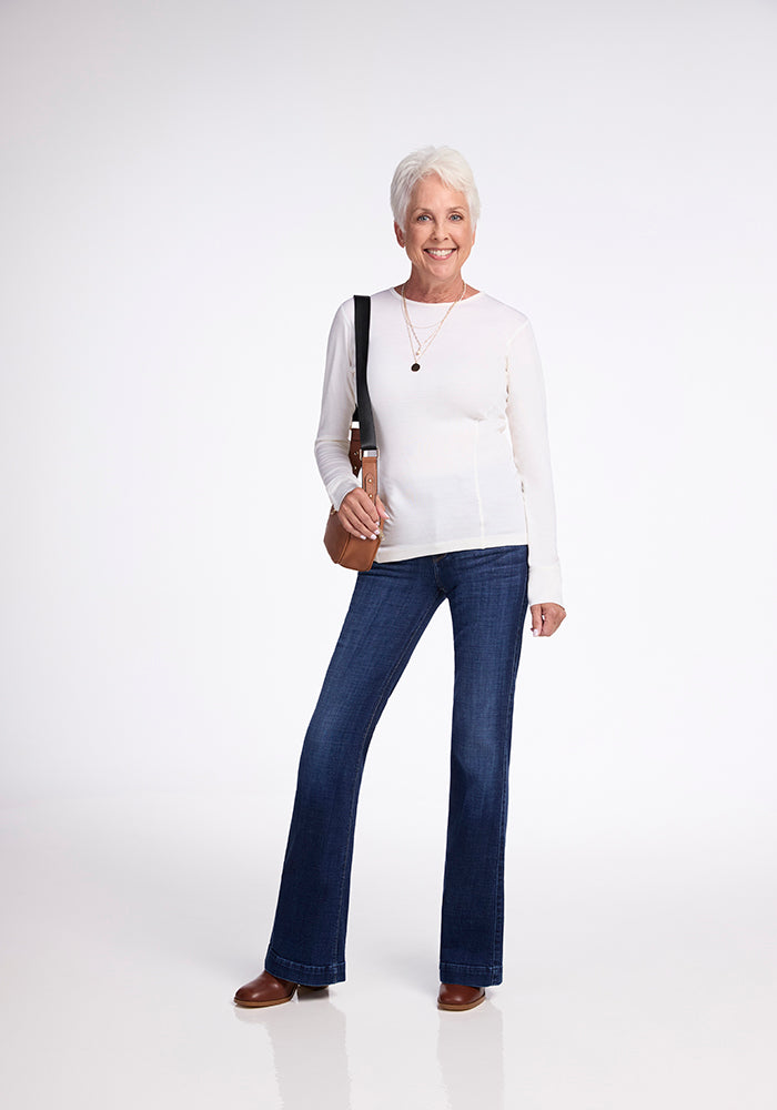 A woman with short white hair smiles against a plain background. She's wearing the Woolx Hannah Long Sleeve Top in Buttercream, blue flared jeans, and brown shoes. She carries a brown leather bag on her shoulder and wears a pendant necklace.
