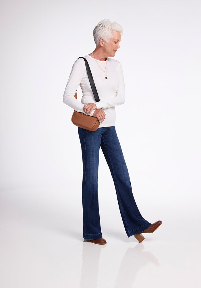 An older woman with short gray hair wears a buttercream Hannah Long Sleeve Top from Woolx and blue flared jeans. She carries a brown bag over her shoulder, pairs it with brown shoes, and walks while looking to the side against a plain background.