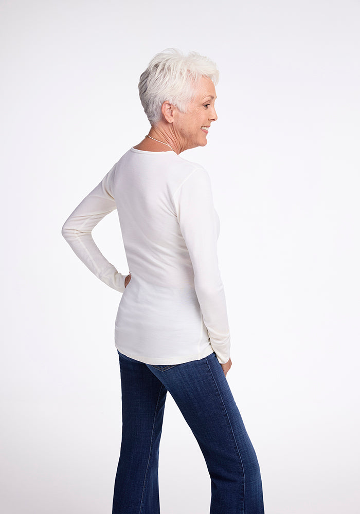 An older woman with short white hair smiles and looks to the side, wearing a long-sleeved Hannah Top by Woolx in buttercream and blue jeans against a plain white background.