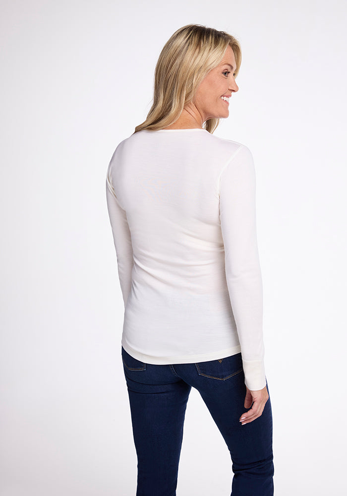 A woman with long blonde hair stands, smiling slightly to the right. She wears a sleek Woolx Hannah Long Sleeve Top in Buttercream and dark blue jeans against a plain white background.