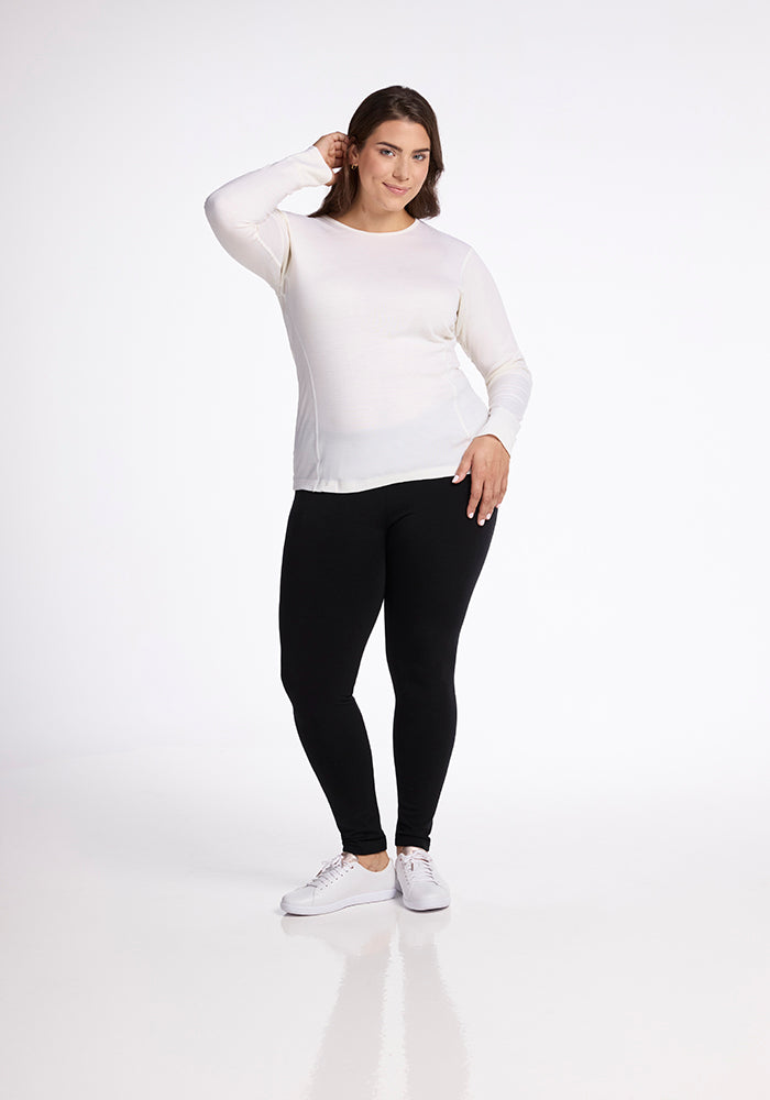 Against a white background, a confident person wears the moisture-wicking Hannah Long Sleeve Top in Buttercream by Woolx, along with black leggings and white sneakers. They have medium-length brown hair and are softly smiling.