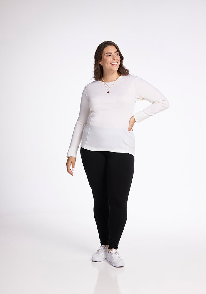 A person stands against a plain white background, smiling. They wear a Hannah Long Sleeve Top in buttercream from Woolx, black pants, and white sneakers. With long hair and one hand on their hip, they pose confidently.