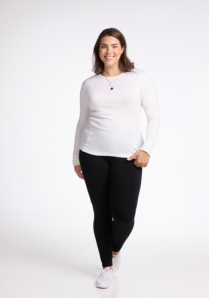 A woman with long brown hair smiles as she wears the Woolx Hannah Long Sleeve Top in buttercream, paired with black leggings and white sneakers. She stands against a light background, holding the bottom of her top with one hand and wearing a necklace.