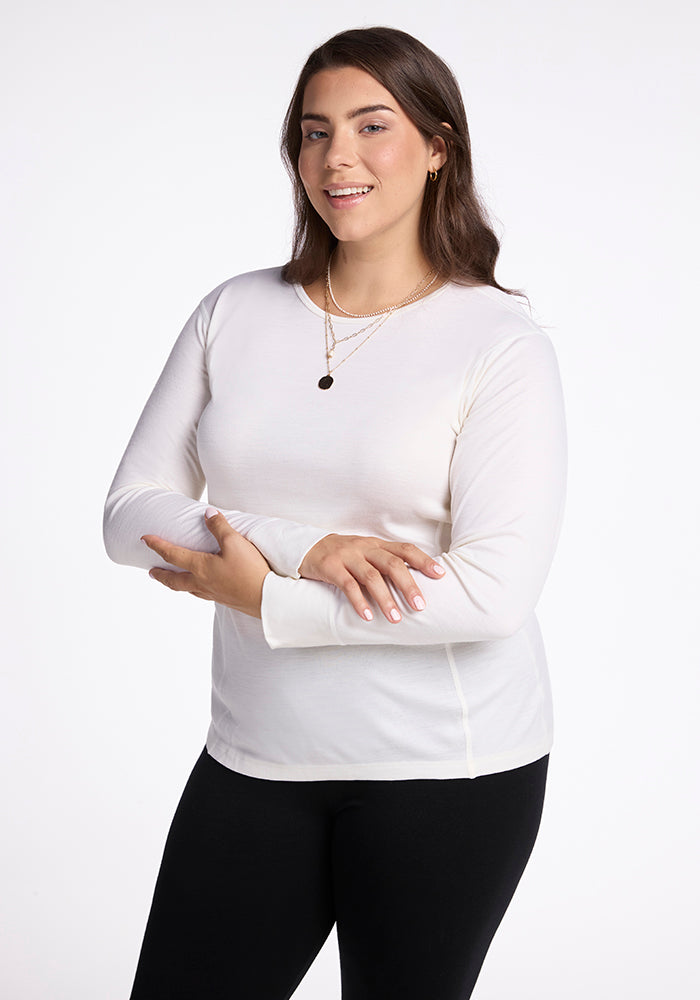 A woman with long brown hair wearing a Woolx Hannah Long Sleeve Top in buttercream and black pants. She accessorizes with gold necklaces and poses with her arms crossed against a white background. 