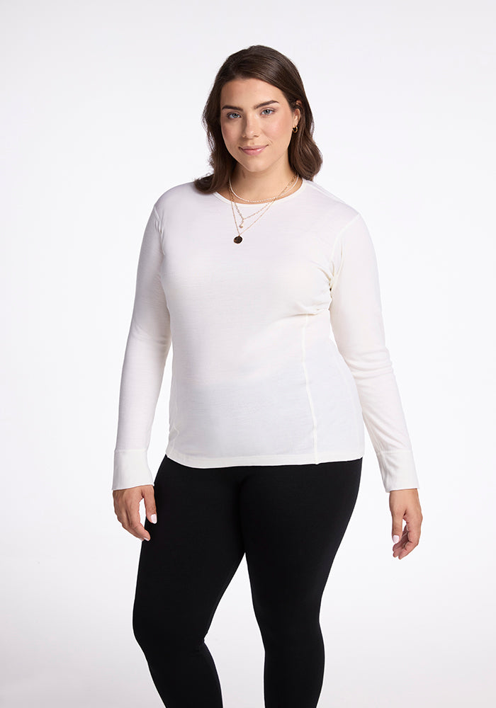 A woman with long brown hair is wearing the Hannah Long Sleeve Top in Buttercream from Woolx paired with black pants. She's accessorized with two necklaces and smiles softly at the camera against a plain white background.