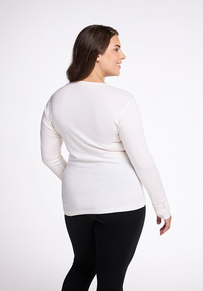 A woman, with her back to the camera, wears a Woolx Hannah Long Sleeve Top in buttercream and black pants. Ideal as a base layer, her outfit harmonizes with the plain white background as she faces slightly left.