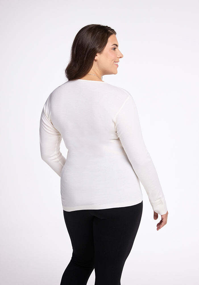 A person with long brown hair stands with their back to the camera, wearing the Woolx Hannah Long Sleeve Top in Buttercream and black pants. Ideal for moisture-wicking, they pose against a plain white background.