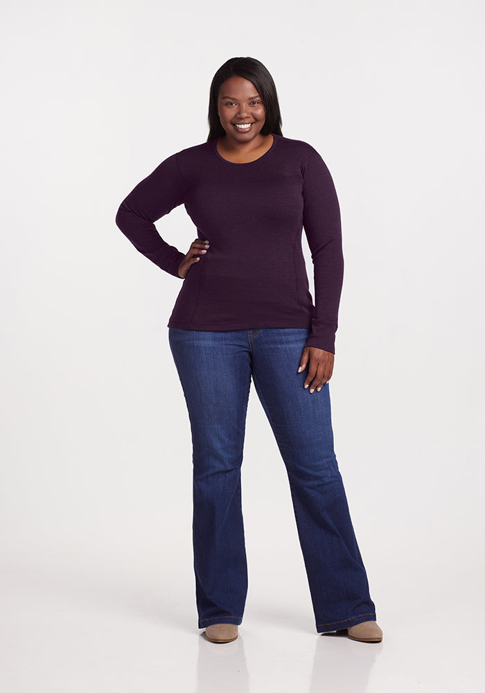 A woman smiles, wearing the Woolx Hannah Long Sleeve Top in Deep Plum, styled with blue jeans. Her left hand is on her hip, right arm relaxed, and she's sporting beige shoes against a plain white background.