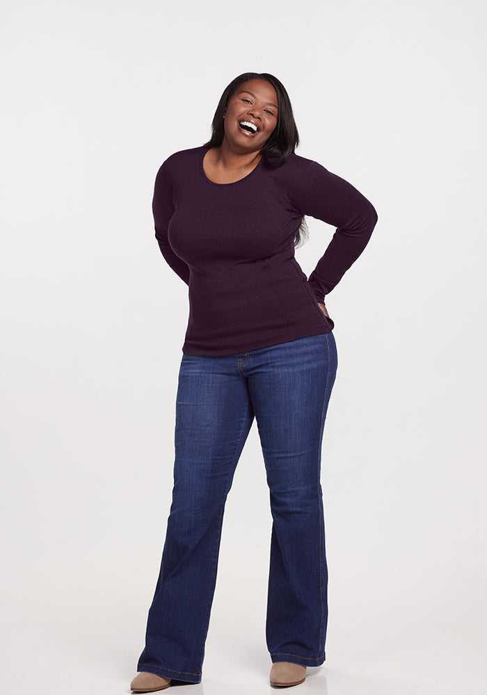 A person smiling, standing with hands behind their back, wears a Woolx Hannah Long Sleeve Top in Deep Plum, paired with blue jeans and brown shoes against a plain white background.