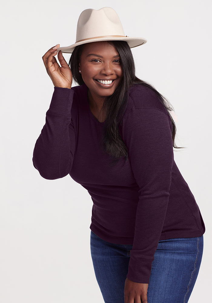A smiling woman with long dark hair wears a cream-colored hat, the Woolx Hannah Long Sleeve Top - Deep Plum, and blue jeans. She playfully tilts the hat with one hand while posing against a neutral background. 