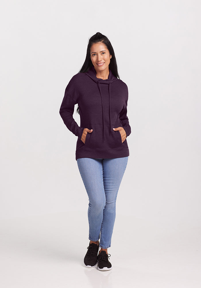 A long-haired person smiles, wearing a Woolx Callie Hoodie in Deep Plum, paired with light blue jeans and black sneakers. They stand against a plain white background.