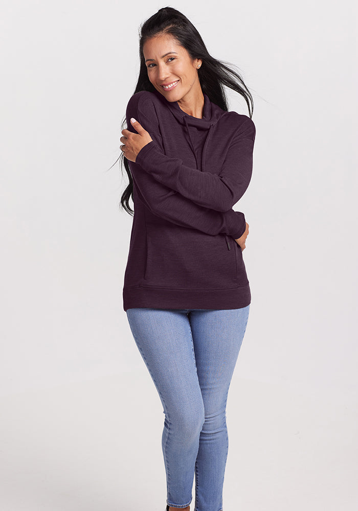 A person stands smiling, wearing a cozy Callie Hoodie in Deep Plum with light blue jeans. With long dark hair and arms crossed, they pose against a white background, showcasing this Woolx layering essential.
