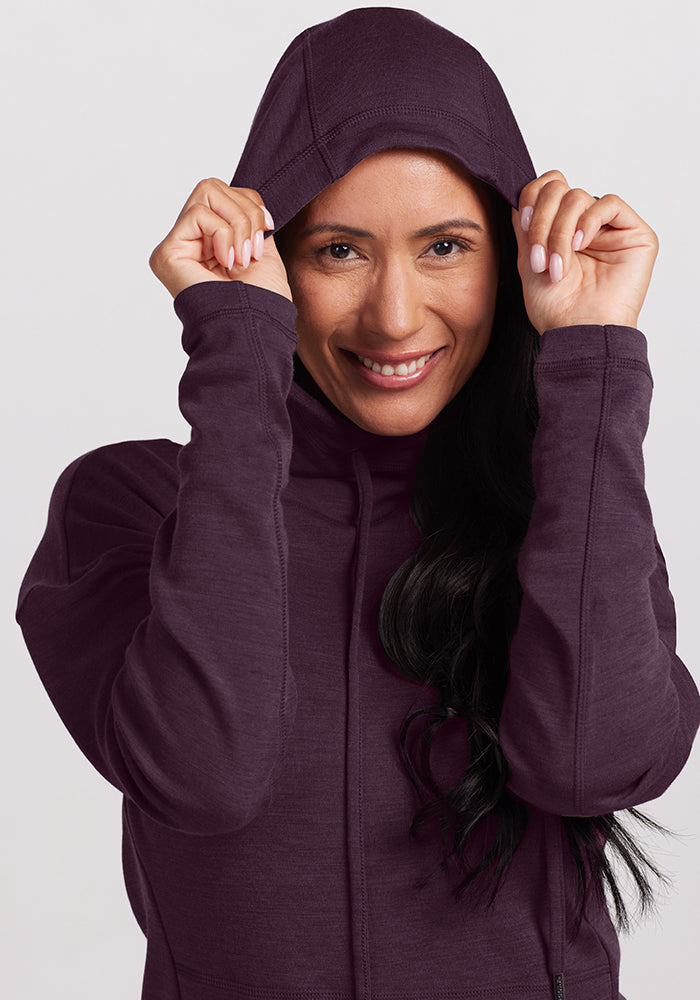 Smiling, they pull the hood of their Woolx Callie Hoodie in Deep Plum over their head, snugly layering it over long sleeves and dark hair against a light background. Final Sale Deals!.