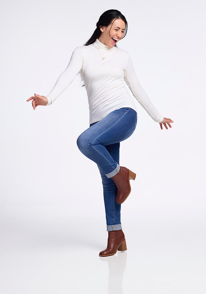 Sporting a Buttercream Peyton Turtleneck from Woolx, blue jeans, and brown heeled ankle boots, a person stands on one leg with a smile, playfully extending their arms against a plain, light background.