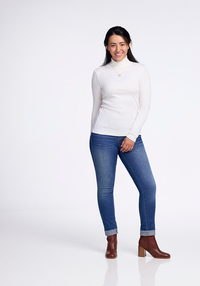 A woman stands smiling against a white background, wearing the Woolx Peyton Turtleneck in Buttercream, paired with blue jeans and brown heeled boots. Her dark hair is down, with her hands relaxed at her sides.